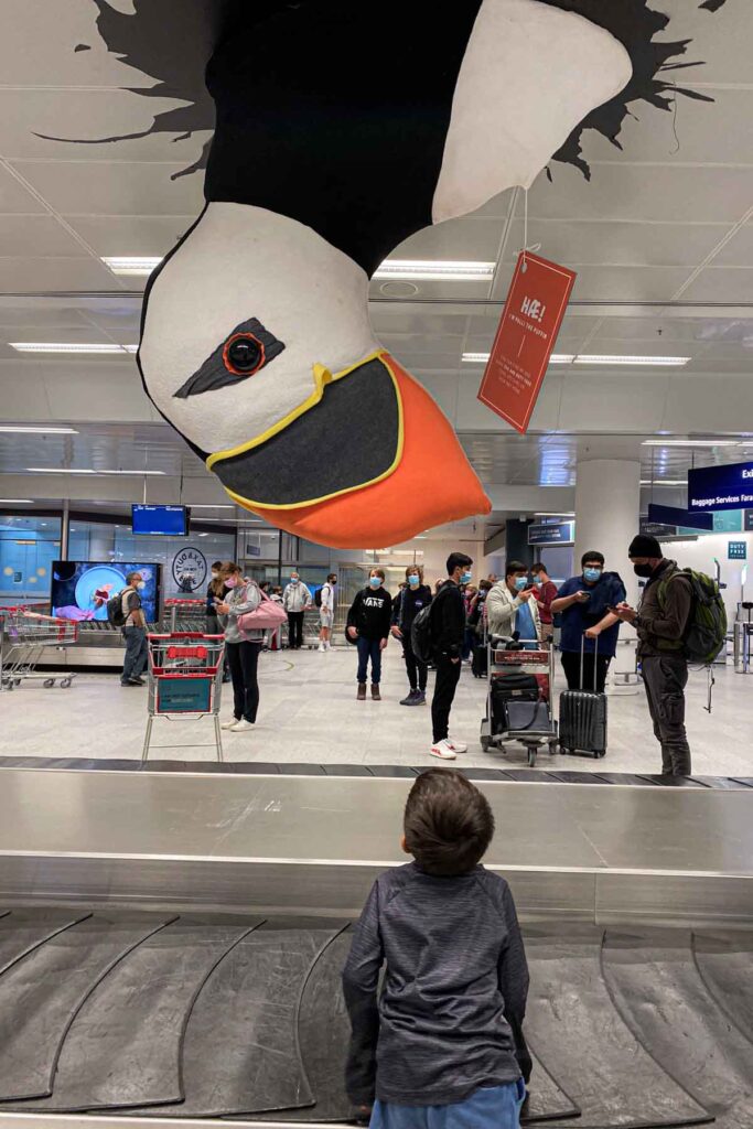 A young child stares up in awe at a giant puffin sculpture hanging above a baggage claim belt in the KEF Keflavík International Airport in Iceland, with travelers with suitcases and carts in the background.