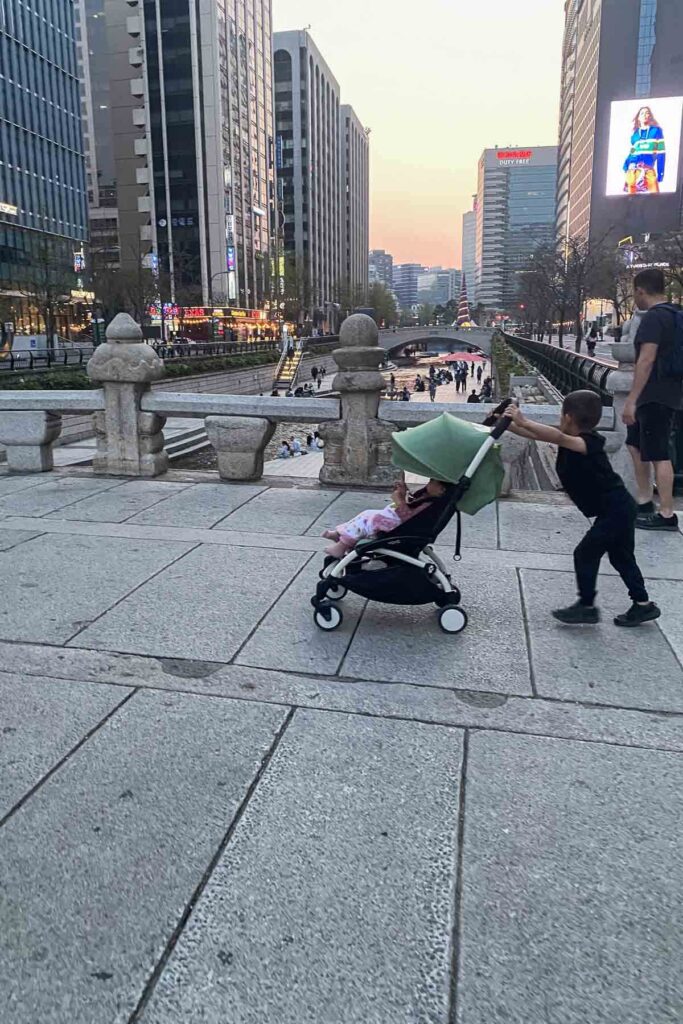 A young boy pushes a stroller with a toddler along a wide stone bridge overlooking Seoul’s Cheonggyecheon Stream, framed by modern skyscrapers and evening city lights.