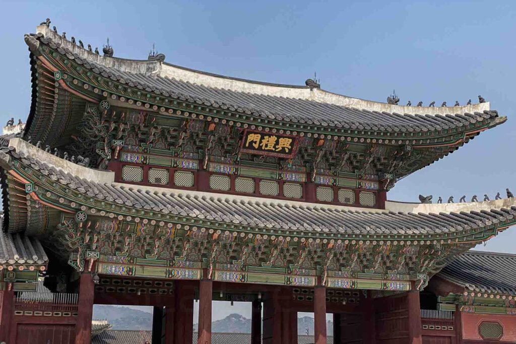 A close-up view of the elaborately decorated roof and wooden eaves of a traditional Korean Gyeongbokgung palace gate in Seoul, featuring vibrant colors, symbolic ornaments, and a golden sign in Hangul.