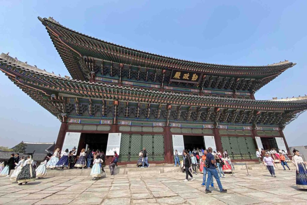 A wide view of the intricately detailed Geunjeongjeon Hall at Gyeongbokgung Palace, with tourists—many in colorful hanbok—gathered around the grand entrance on large flat stones in Seoul in spring.