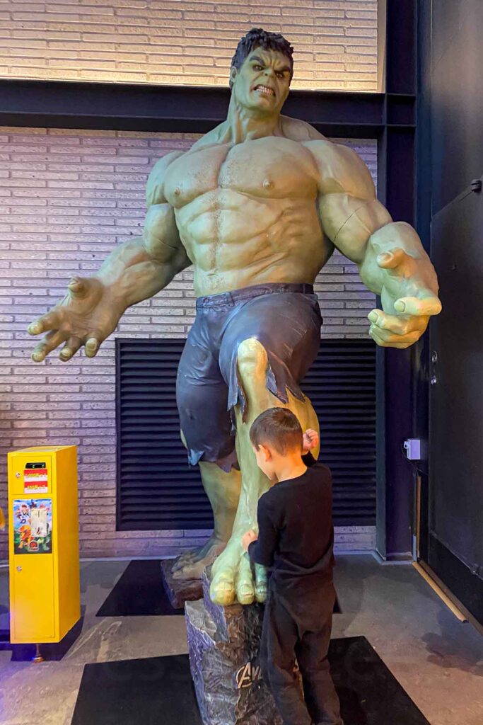 A young boy wearing black mimics a strongman pose next to a towering, muscular Hulk statue in torn pants at a themed Marvel indoor display area in IPark Mall, Seoul.