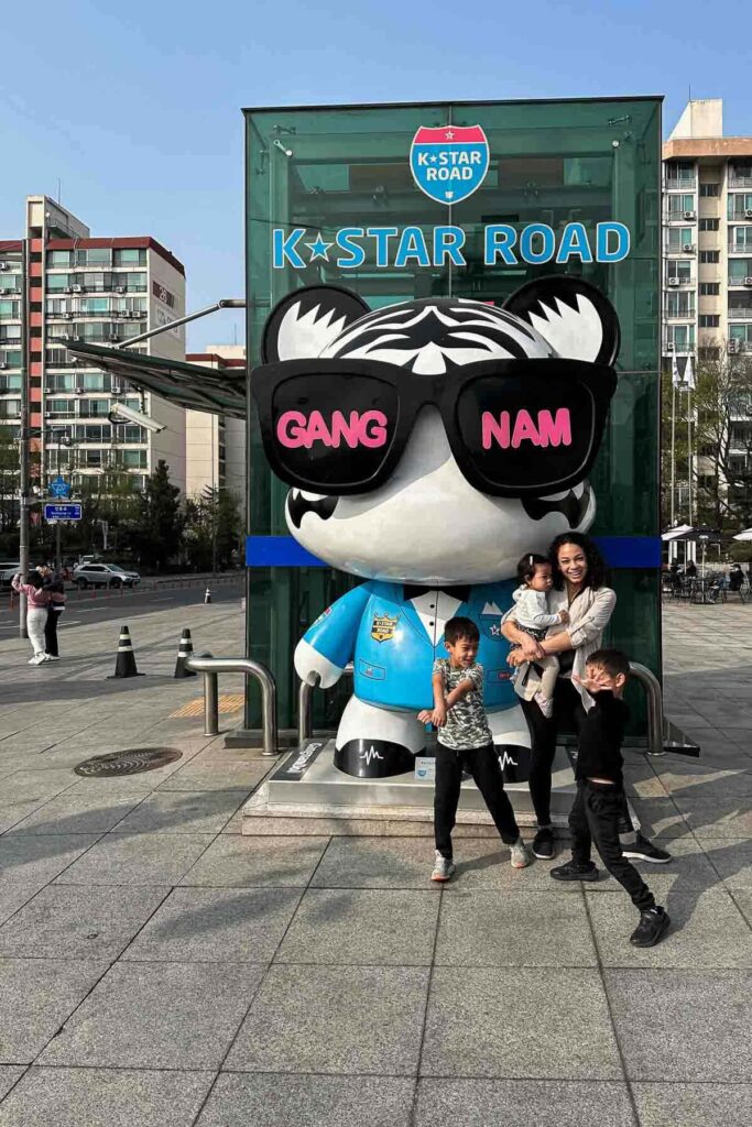 A mom holding a toddler poses with two boys in front of a giant bear statue wearing sunglasses labeled “GANG NAM” at the K-Star Road landmark in Gangnam, Seoul, with high-rise buildings in the background.