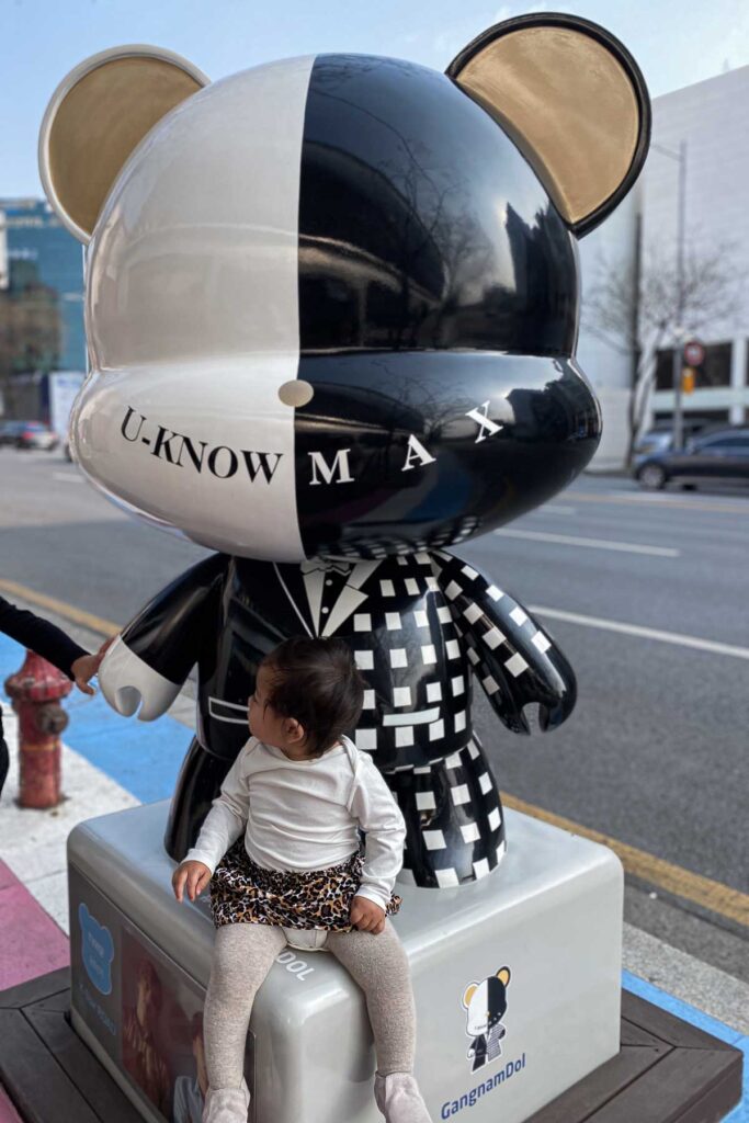 A toddler in a leopard skirt sits on a black-and-white checkered Gangnamdol bear statue labeled “U-KNOW MAX” on a sidewalk in Seoul.