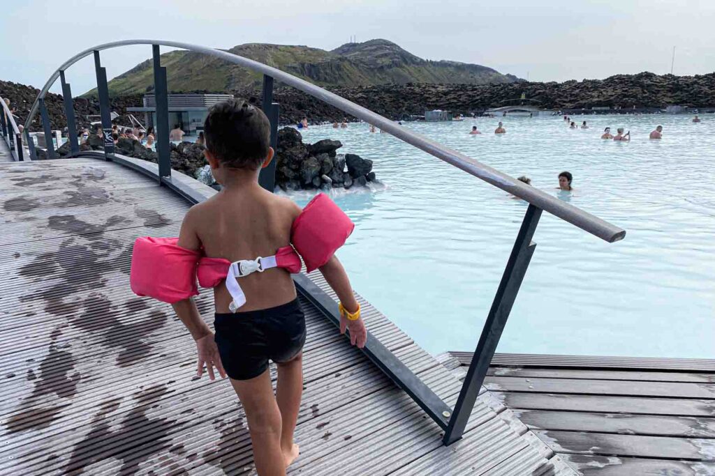A child wearing pink puddle jumper walks barefoot up an arch wooden bridge over the Blue Lagoon, heading toward a calm area of the water where others are swimming, surrounded by dark volcanic rock walls.