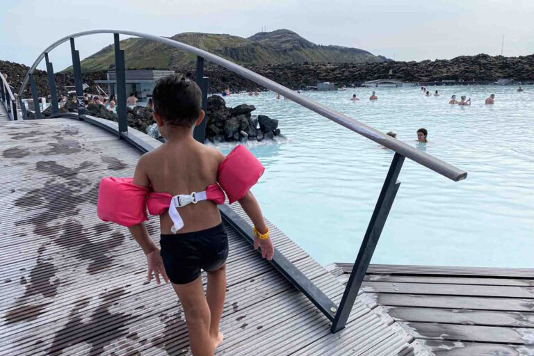 A child wearing pink puddle jumper walks barefoot up an arch wooden bridge over the Blue Lagoon, heading toward a calm area of the water where others are swimming, surrounded by dark volcanic rock walls.