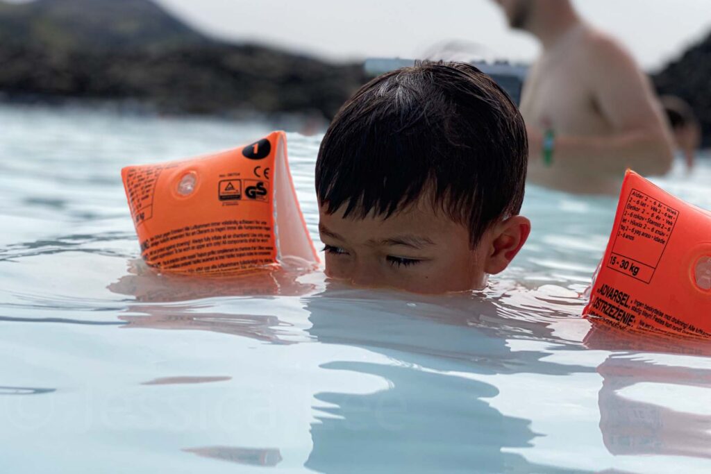Close-up of a child wearing bright orange floaties, eyes just above the surface of the Blue Lagoon’s mineral-rich water as his nose and mouth are underwater.