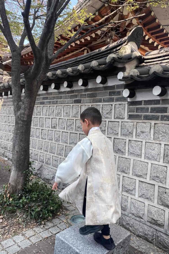 A young boy wearing a cream-colored hanbok stands on a stone block near a traditional Korean stone wall and tiled roof, next to a tree outside Changdeokgung.