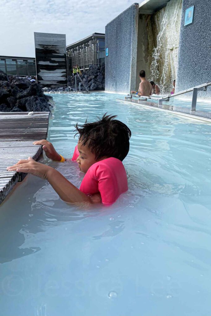 A young child in pink floaties holds the wooden edge of the Blue Lagoon, with a waterfall feature and spa guests relaxing in the background.