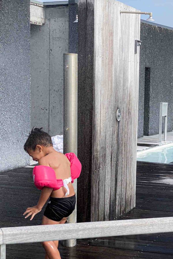 Child wearing bright pink puddle jumper stands under an outdoor shower at the Blue Lagoon on the deck, water streaming down over their back and shoulders.