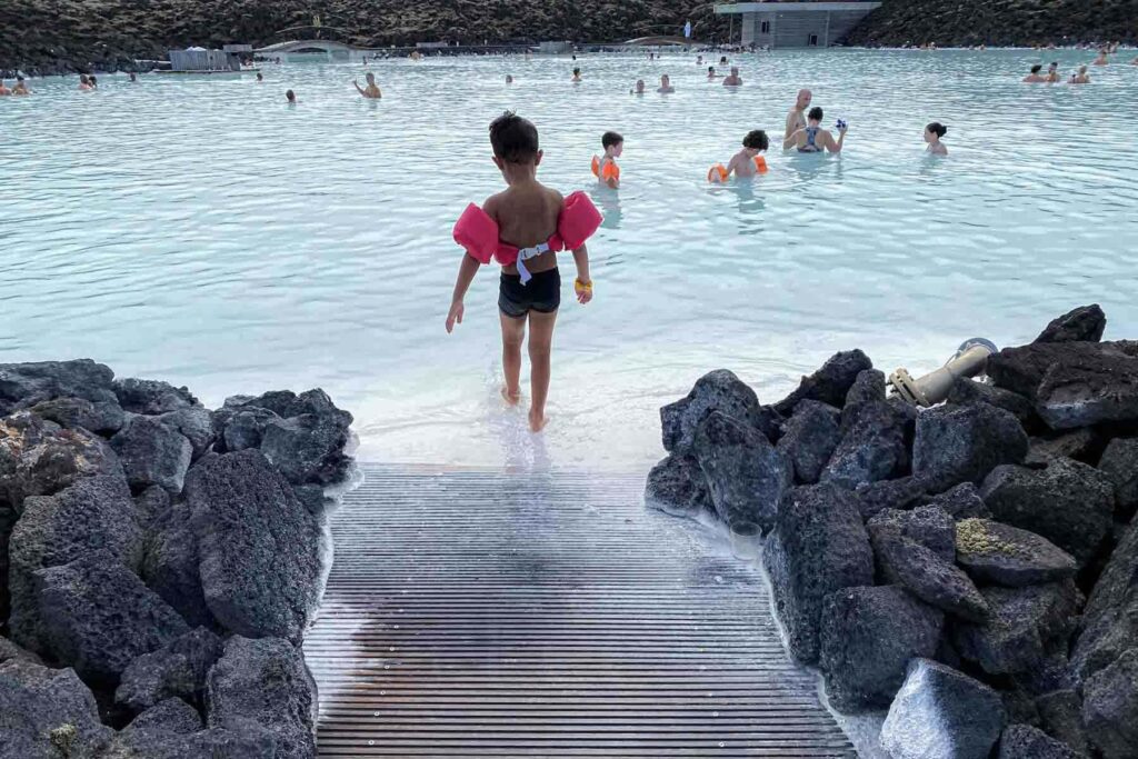 A young child in a pink puddle jumper floatie steps into the shallow edge of the Blue Lagoon, surrounded by black volcanic rocks, as other visitors, including more kids in floaties enjoy the warm geothermal waters.