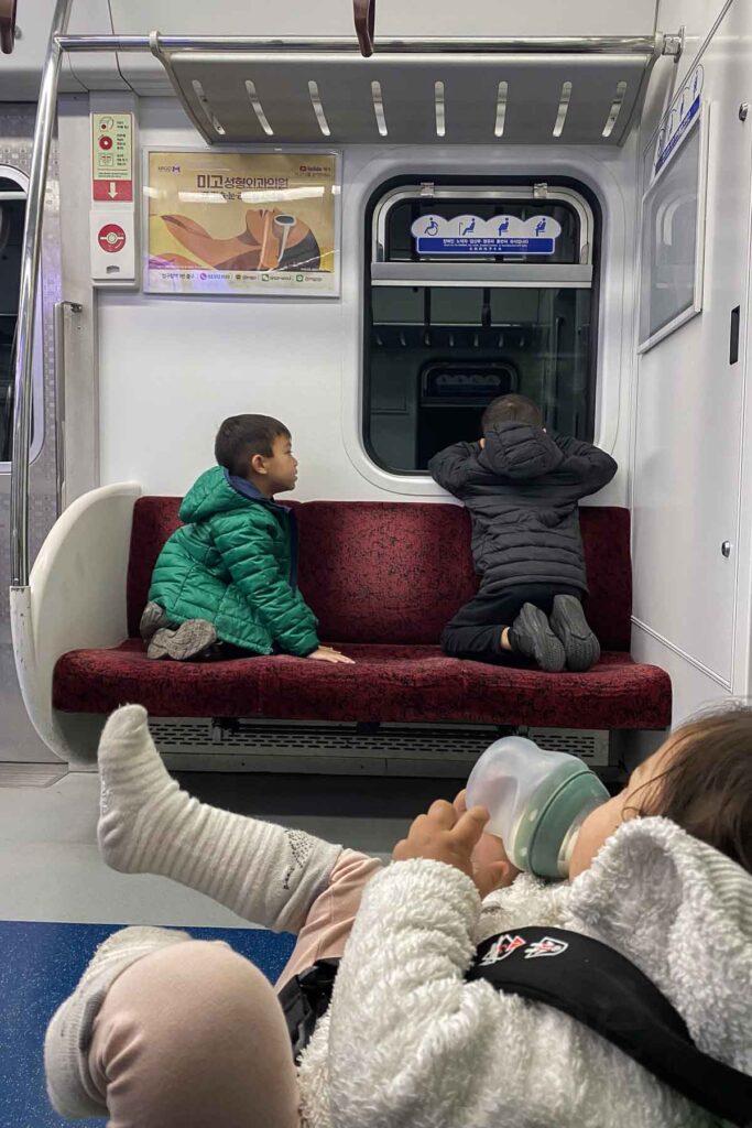 Two children kneel on a deep red subway bench, one looking out the window and the other chatting, while a baby in a stroller wearing a fuzzy white coat drinks milk from a bottle in the foreground.