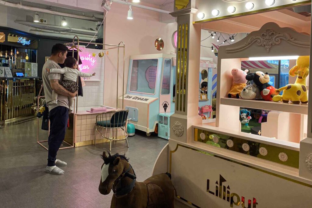A father holds his toddler who reaches toward a pastel pink beauty play station inside a children's Lilliput play café in Seoul filled with posh and plush toys and pretend setups.