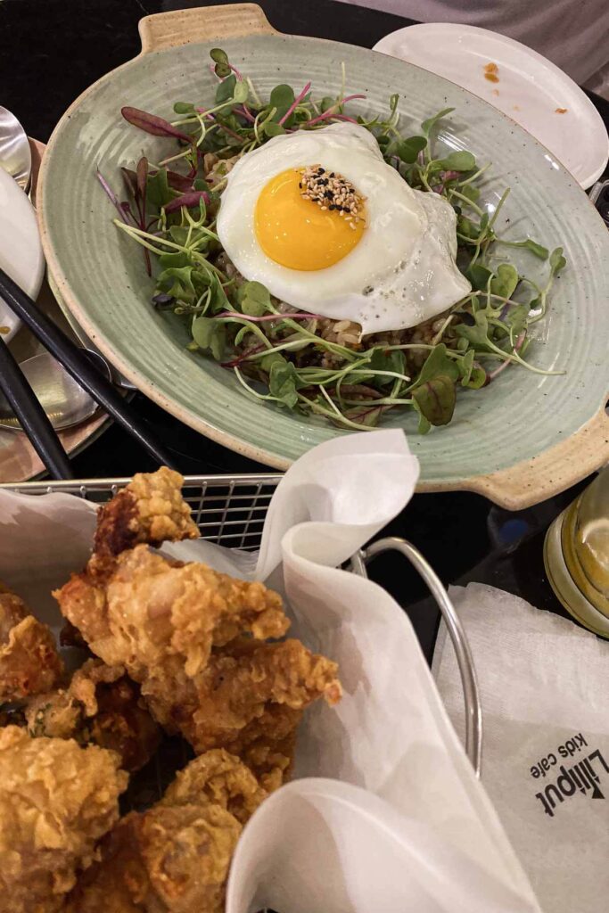 A bowl of stir-fried rice topped with greens and a sunny-side-up egg garnished with sesame seeds, served alongside a basket of crispy Korean fried chicken at the Lilliput Kids Cafe in Seoul.