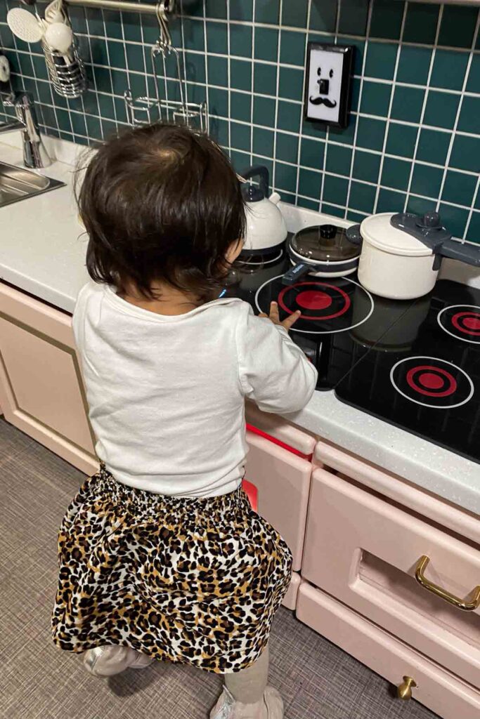 A toddler wearing a leopard-print skirt and white shirt stands at a toy kitchen set, reaching toward a pretend stovetop with plastic pots and pans and lower light pink cabinets at Lilliput Cafe in Seoul.