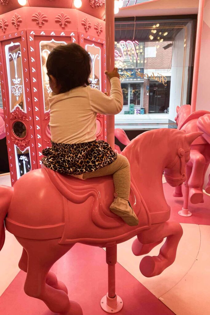 A young toddler girl wearing a leopard print skirt sits on a pink carousel horse inside a whimsical indoor amusement area decorated in all pink tones at Lilliput Kids Cafe in Seoul, South Korea.