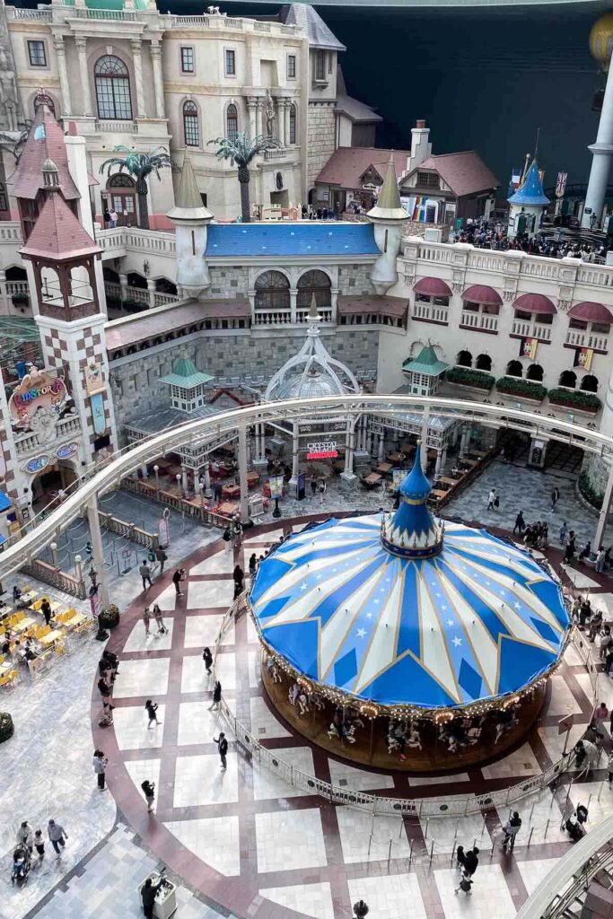 A top-down view of a whimsical indoor theme park with a blue-and-gold carousel at the center, surrounded by castle-style buildings, a monorail track, and visitors walking through the plaza at Lotte World amusement park in Seoul.