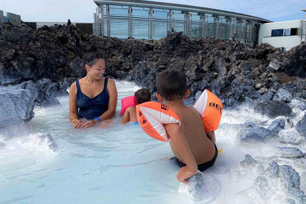 Pregnant mom and two children with floaties relax in a shallow corner of the Blue Lagoon surrounded by jagged black rocks, capturing a peaceful family moment at the Blue Lagoon.