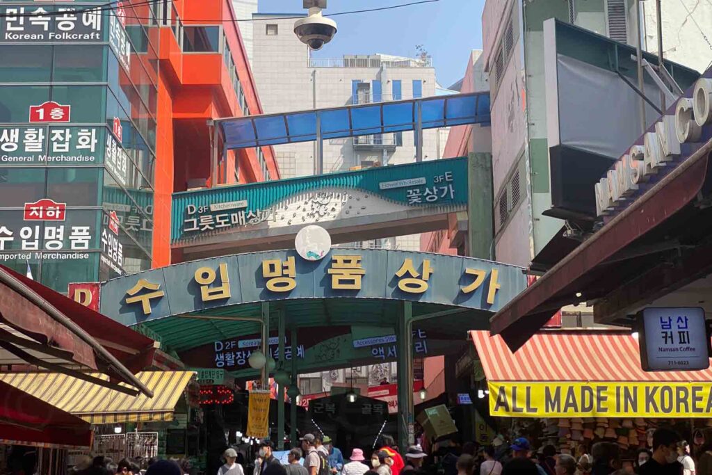 The bustling entrance to a Namdaemun market in Seoul with bold signage in Hangul and English, surrounded by colorful buildings and crowded vendor stalls.