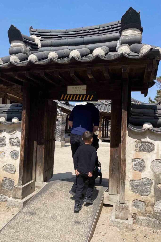 A dad in blue and a child in black walk under a low wooden gate with a tiled roof marked "Watch Your Head," entering a traditional Korean house courtyard in Namsangol Hanok Village in Seoul.