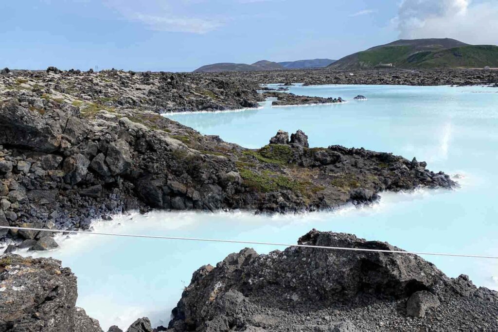 Expansive view of milky blue geothermal pools stretching between black lava fields near the Blue Lagoon, with distant hills and a mostly clear sky in summer.