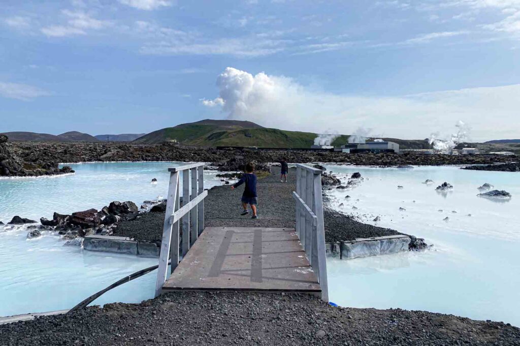 Two children walk across a small wooden bridge over pale blue geothermal water outside the Blue Lagoon, with steam rising in the background from the nearby power plant.