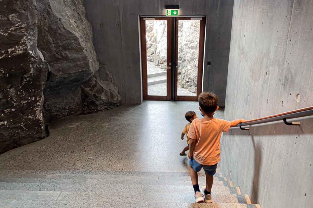 Two young kids walk down a concrete stairwell inside the Blue Lagoon facility, heading toward an exit framed by volcanic rock and glass doors.