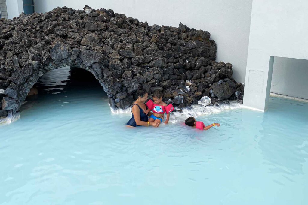 Mother cradles one child as another flots nearby both wearing pink floaties as they stand near a lava rock arch in a shallow section of the Blue Lagoon.