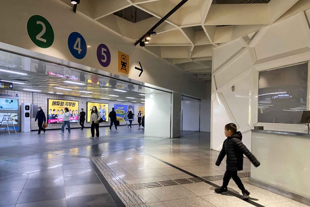A child in a black puffer jacket walks through a bright clean subway corridor toward the direction signs for Seoul Metro lines 2, 4, and 5, surrounded by commuters.