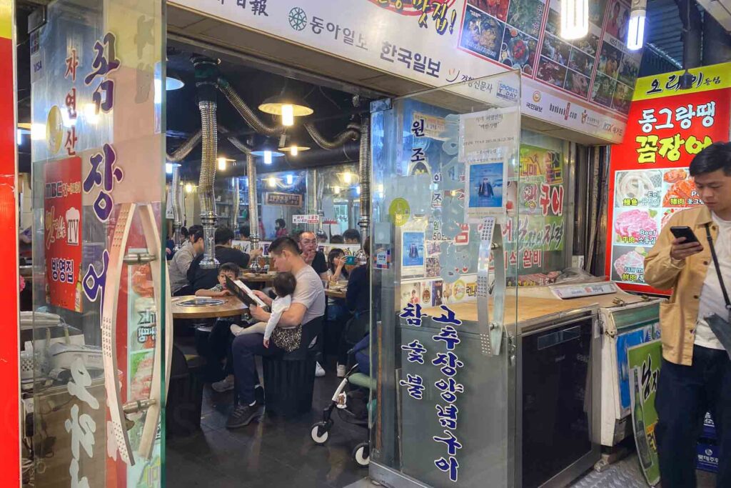 Inside view (from someone standing outside the door) of a busy Korean barbecue restaurant covered in Korean signs and images filled with customers, including a man holding a baby while browsing the menu, under metal ventilation pipes.