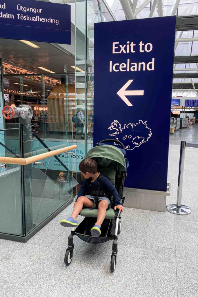 Child in a green yoyo travel stroller waits near a large deep blue airport sign that reads “Exit to Iceland" and a graphic outline of Iceland, with directional arrow pointing left and terminal glass walls in the background.