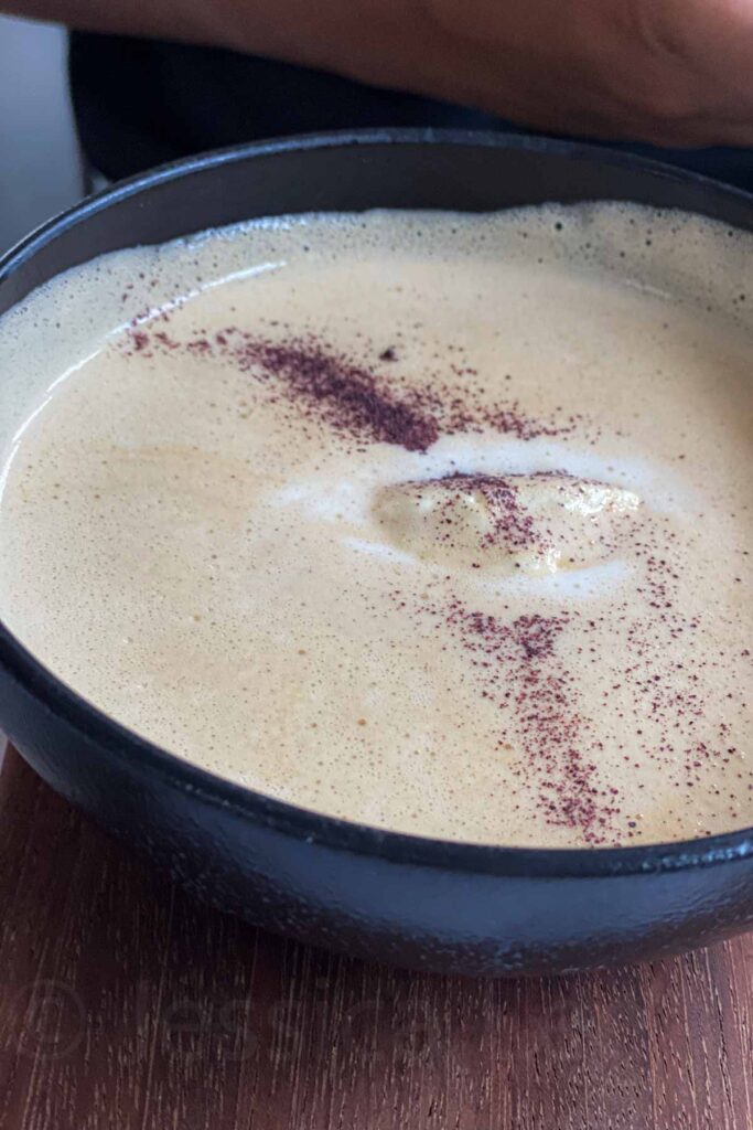 Close-up of a creamy soup served in a black bowl, garnished with fine reddish-brown powder and a swirl of froth.
