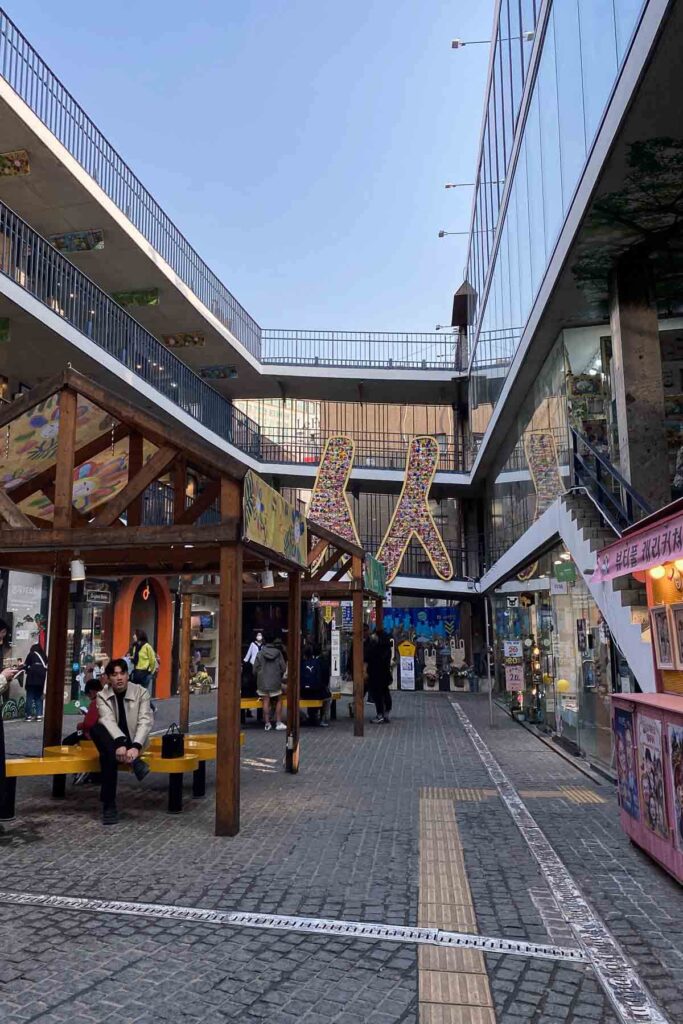 A multilevel open-air shopping center Ssamziegil with glass walls, colorful signage, and outdoor wooden benches, where people are walking and browsing small local stores.