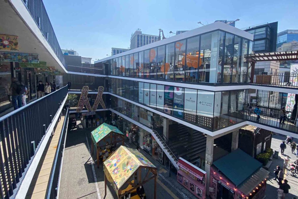 A three-story open-air shopping plaza Ssamziegil with modern glass storefronts, colorful art, and food stalls on the ground level, bustling with visitors in Seoul’s Insadong area.