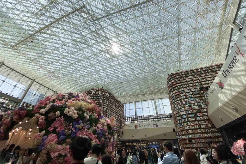 The spacious, glass-ceilinged library atrium of Starfield COEX Library in Seoul, South Korea, filled with people, featuring towering curved bookshelves and a large dome-shaped floral installation made of colorful artificial flowers.