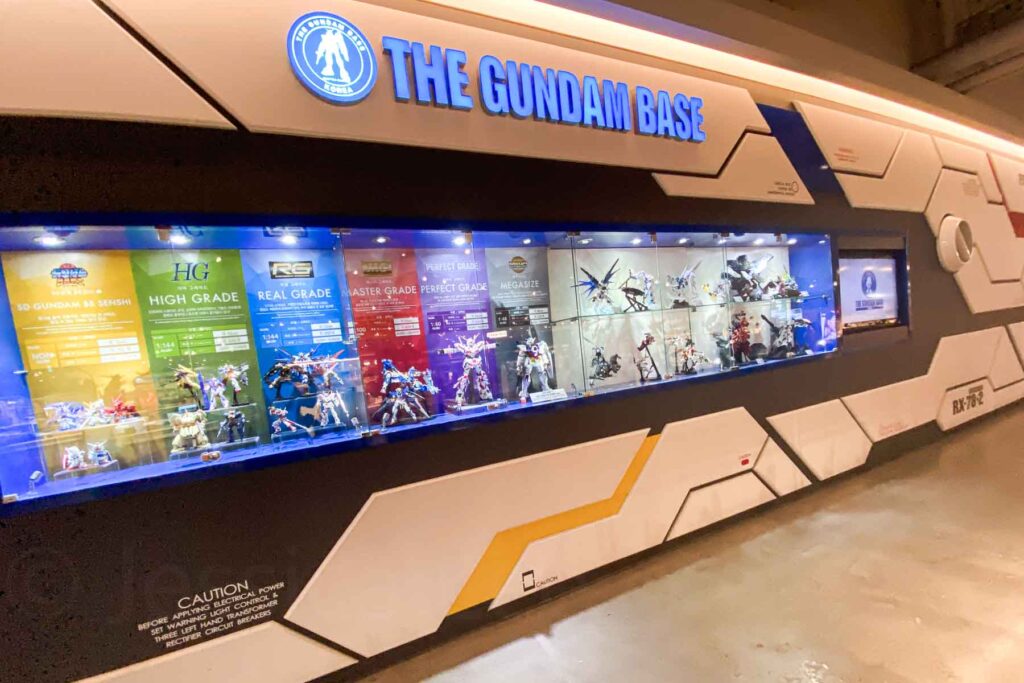 A wide white space-ship-like display boasts the title “The Gundam Base” at the I’Park Mall in Seoul, showing colorful display cases for various grades of Gundam model kits under bright lighting, with informational panels for each series.