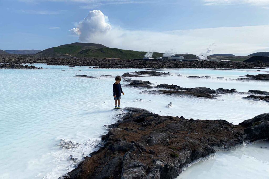 Young child explores rocky terrain along the edge of the Blue Lagoon, standing on a dark lava barely visible above the misty blue geothermal pools.