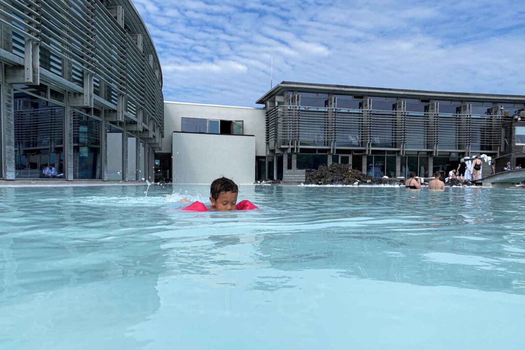 Young child in pink floaties swims in the Blue Lagoon, with the spa’s modern concrete and glass buildings in the background.
