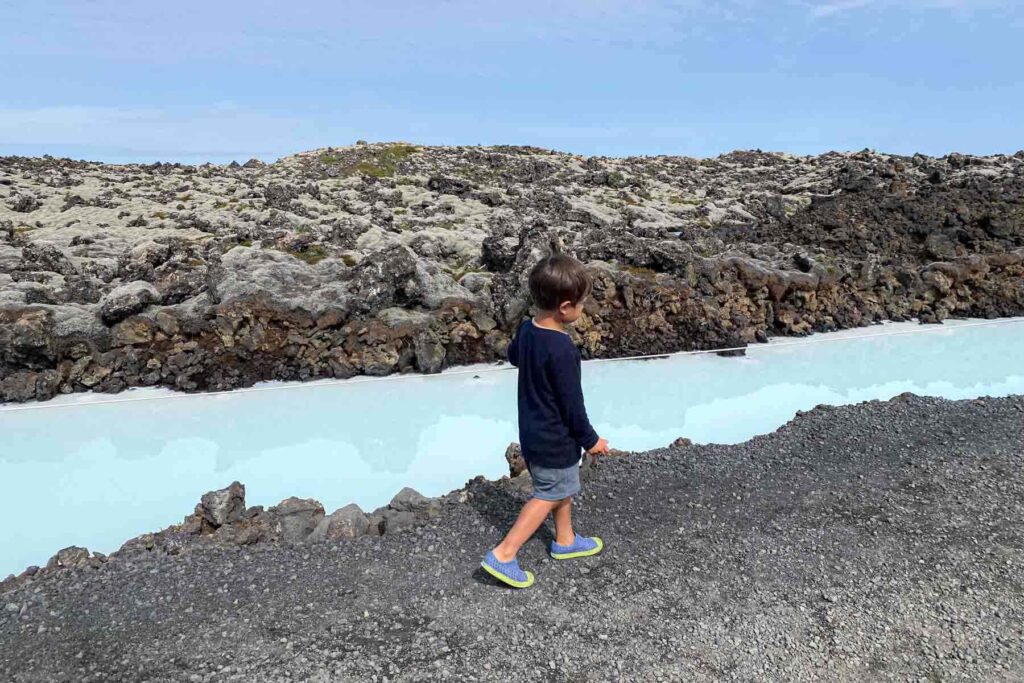Child in a long-sleeve navy shirt walks along a gravel path beside a narrow blue geothermal stream surrounded by black lava rocks near the Blue Lagoon in Iceland.