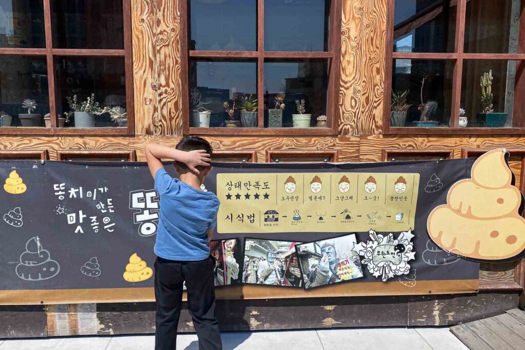 A boy stands with one hand behind his head, looking at a humorous sign covered in cartoon poop illustrations and Korean writing outside a novelty-themed café in Insadong, Seoul.