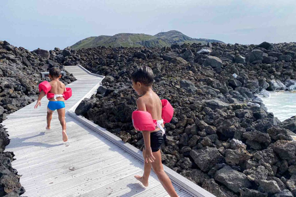 Two young boys in swim trunks wearing bright pink floaties walk along a winding wooden boardwalk through a rugged lava rock landscape at the Blue Lagoon.
