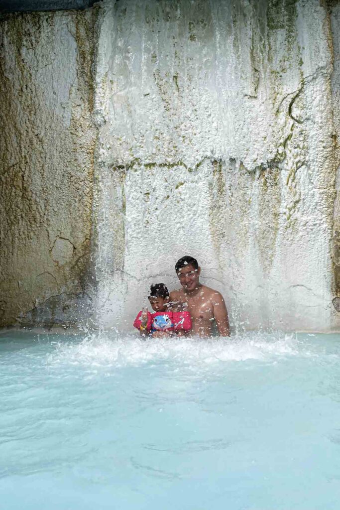 Father and young child in puddle jumper stand beneath a strong waterfall at the Blue Lagoon, with splashes of water surrounding them in the warm pool.