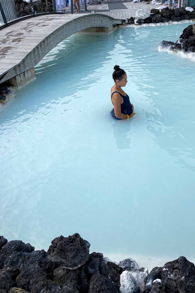 Woman in a navy swimsuit stands quietly holding her small pregnant belly in a secluded section of the Blue Lagoon, near a wooden bridge and surrounded by black lava rocks and pale blue water.