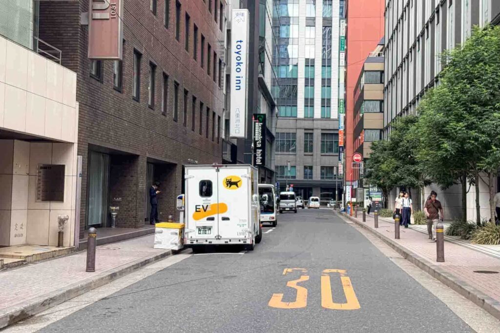 A Yamato Transport electric delivery van is parked on a quiet city street in Tokyo, Japan, with "30" painted on the road and pedestrians walking along the sidewalk. It’s parked below a vertical Tokyo Inn sign, likely doing the daily drop off and pickup.