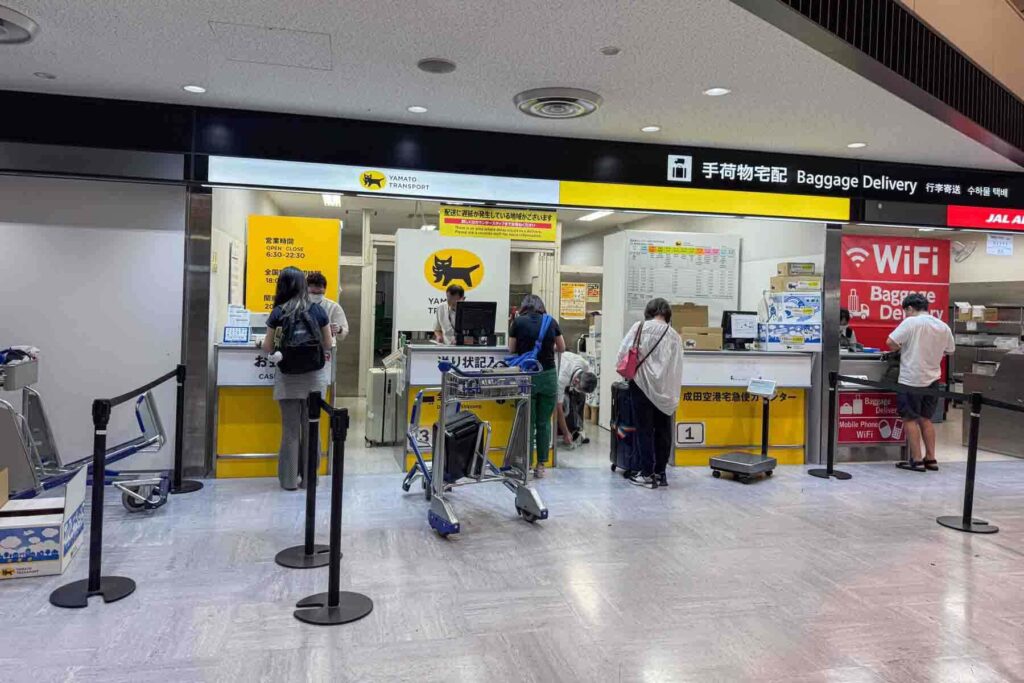 Travelers queue at a Yamato Transport baggage delivery counter with bright yellow signage inside Japan’s NRT airport, surrounded by luggage carts and informational signage. On the side is red signage of another baggage services company.