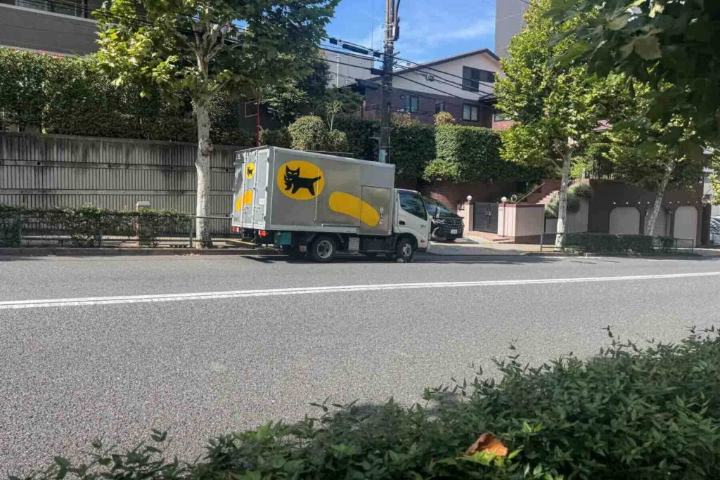A silver Yamato Transport truck with the iconic yellow and black cat and kitten logo is parked on a leafy residential street in Japan.