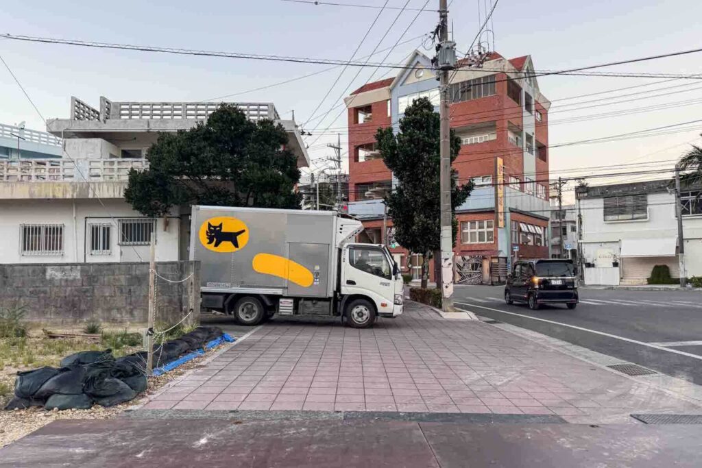 A silver Yamato truck with the iconic black cat logo is parked near a quiet Japanese intersection in Ishigaki, Okinawa, highlighting the far stretches of suitcase delivery service in Japan.