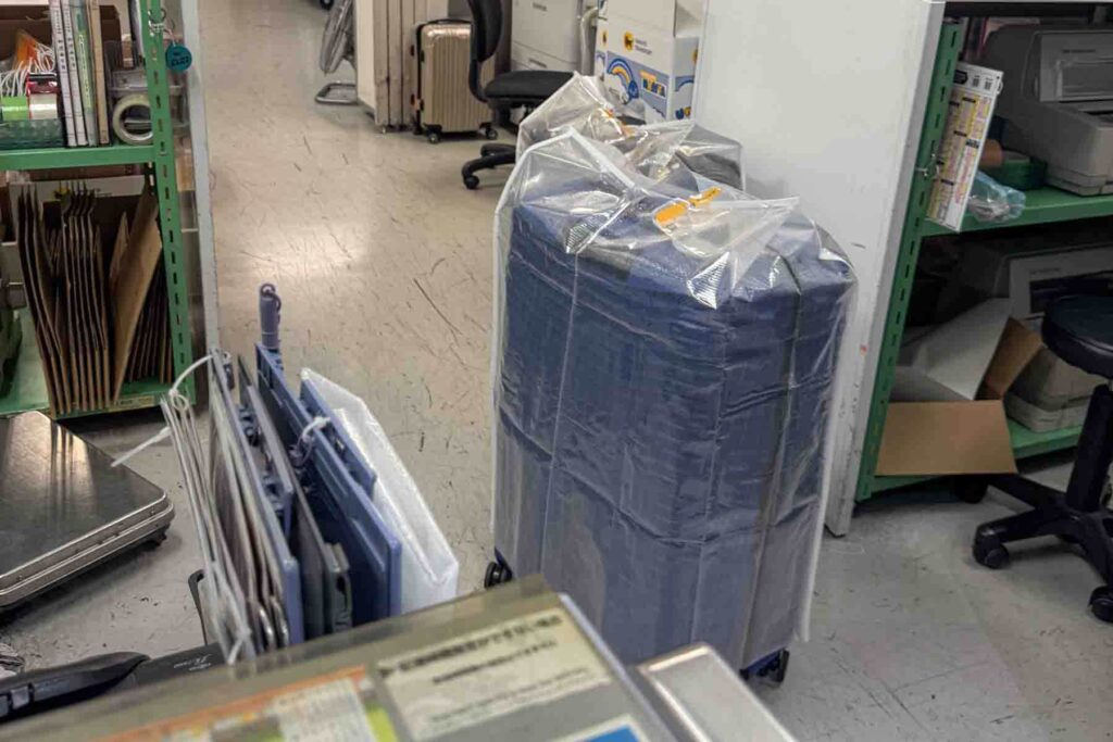 A large blue suitcase fully wrapped in a thick plastic cover with a Yamato tag is staged in a shipping office, surrounded by packaging supplies and office shelves. 
