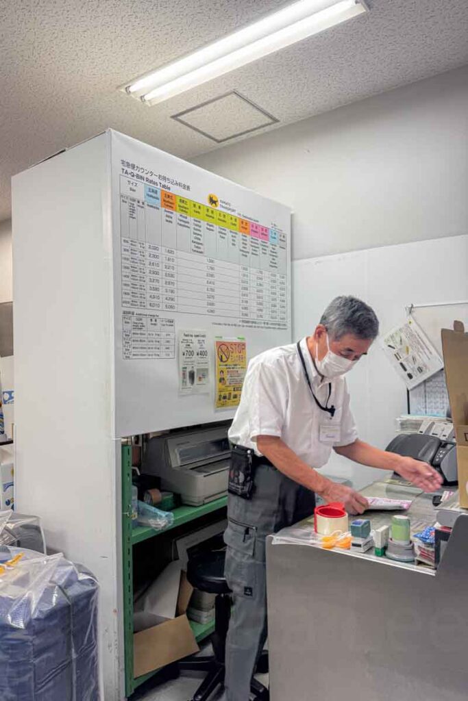 A Yamato staff member in uniform fills out a shipping label beside a wall-mounted rate chart and various packaging tools, like tape and stamps. The image shows the hands-on support travelers receive when using Yamato luggage transfer services.