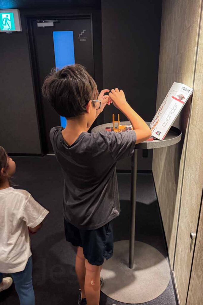 A young boy wearing glasses plays the board game Quoridor in the elevator hall at a hotel in Osaka, showing the interactive amenities for kids. His younger sister is watching along.
