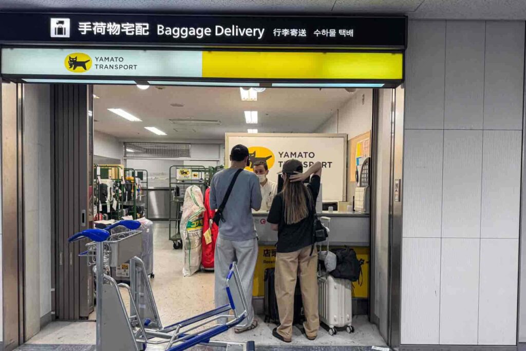 A couple speaks to an agent at the Yamato Transport counter labeled "Baggage Delivery" inside the Haneda airport terminal. They have suitcases by them ready to drop off and an empty luggage cart behind them.